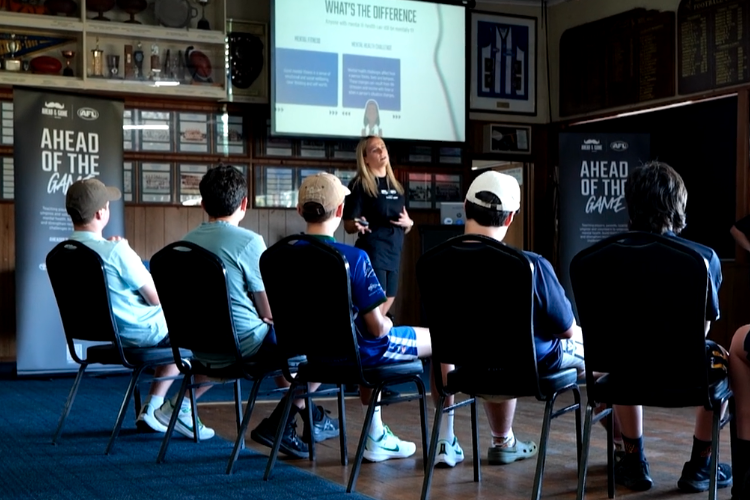 A group of young male kids in sitting in a room with a teacher in from and a projector